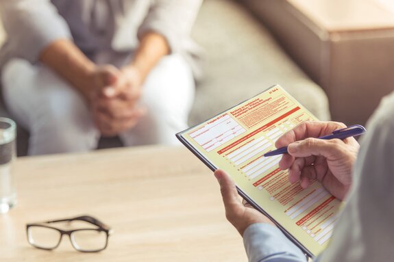 Arzt füllt ein Formular aus. Patient sitzt schemenhaft  im Hintergrund.