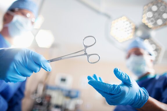 Arzt/Ärztin in blauer OP-Kleidung übergibt eine Schere an Arzt/Ärztin in blauer OP-Kleidung in einem OP-Raum.