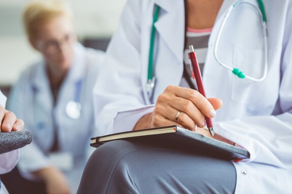 Sitzender Oberkörper einer Ärztin in Arztkittel. Sie hält Notizbuch und Stift auf den Knien. Im Hintergrund unscharf eine zweite Ärztin.