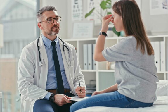 Arzt sitzt auf einem Hocker vor junger Patientin auf einer Liege. Patientin sitzt auf der Liege und zeigt dem Arzt, dass es ihr zwischen den Augen am Kopf weh tut.