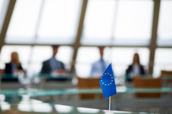 Konferenzraum mit unscharfen sitzenden Menschen im Hintergrund. Im Vordergrund eine kleine stehende EU-Flagge auf einem Tisch.
