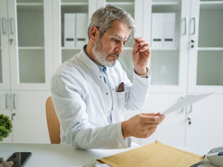 Arzt mit hochgeschobener Brille liest Rechnung am Schreibtisch
