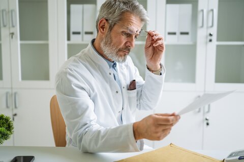 Arzt mit hochgeschobener Brille liest Rechnung am Schreibtisch
