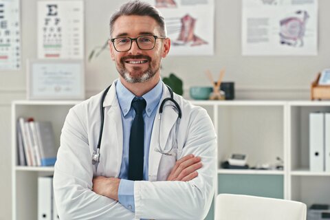 Arzt steht mit verschränkten Armen vor dem Schreibtisch in seiner Praxis