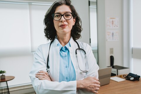 Ärztin steht mit verschränkten Armen vor dem Schreibtisch