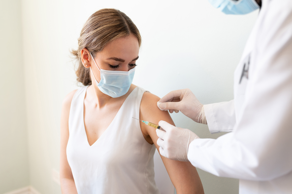 Arzt bereitet den Arm einer Patientin für eine Impfung vor. In der einen Hand hält er einen Tupfer zur Desinfektion, in der anderen die Spritze mit dem Serum. Patientin und Arzt tragen Masken.