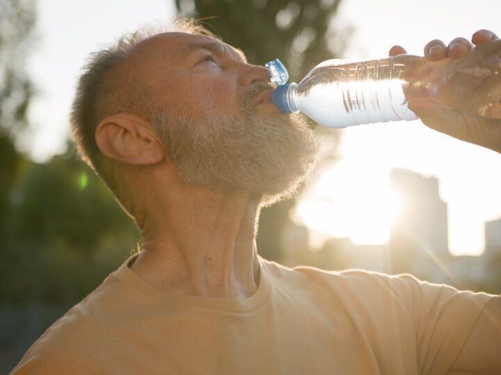 Mann trinkt Mineralwasser aus einer Flasche. Wird von der Sonne angestrahlt.