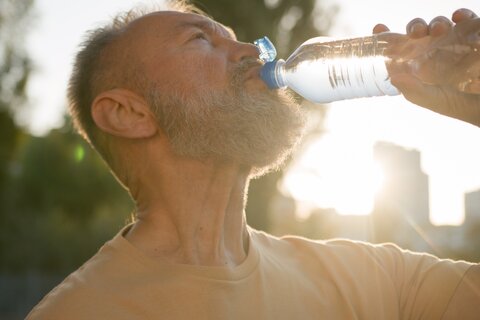 Mann trinkt Mineralwasser aus einer Flasche. Wird von der Sonne angestrahlt.