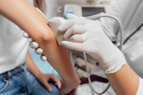 Arzt führt Ultraschalluntersuchung durch. Er führt den Schallkopf des Ultraschallgerätes am Ellenbogen der Patientin entlang.