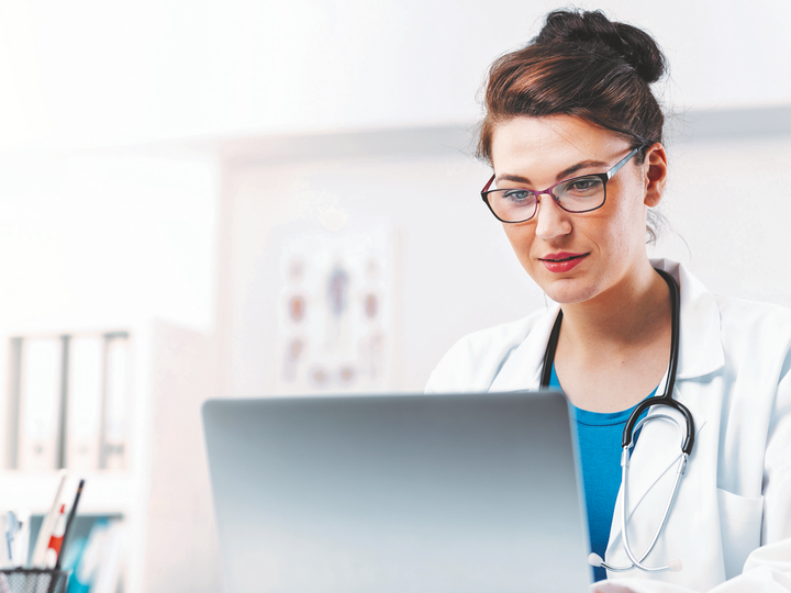 Ärztin mit dunklen Haaren und Brille sitzt an einem Schreibtisch vor einem Laptop.