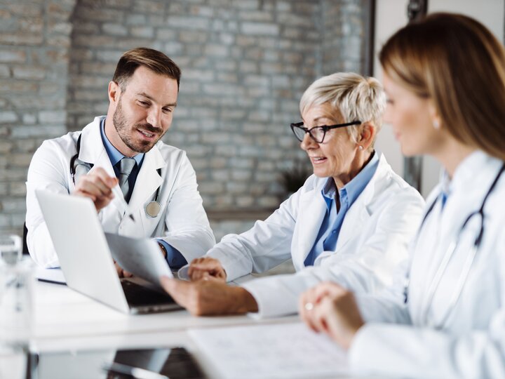 Ein Arzt  in weißem Kittel sitzt mit 2 Kolleginnen an einem Tisch. Vor Ihnen ein aufgeklappter Laptop. Sie unterhalten sich angeregt über ein Dokument, welche eine der Ärztinnen in der Hand hält.