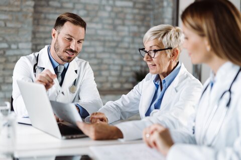 Ein Arzt  in weißem Kittel sitzt mit 2 Kolleginnen an einem Tisch. Vor Ihnen ein aufgeklappter Laptop. Sie unterhalten sich angeregt über ein Dokument, welche eine der Ärztinnen in der Hand hält.