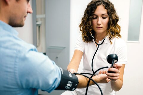 Ärztin mit halblangen, dunklen, lockigen Haaren in weißem Shirt und Hose misst Blutdruck bei einem Patienten mit dunklem kurzen Haar und blauem Hemd