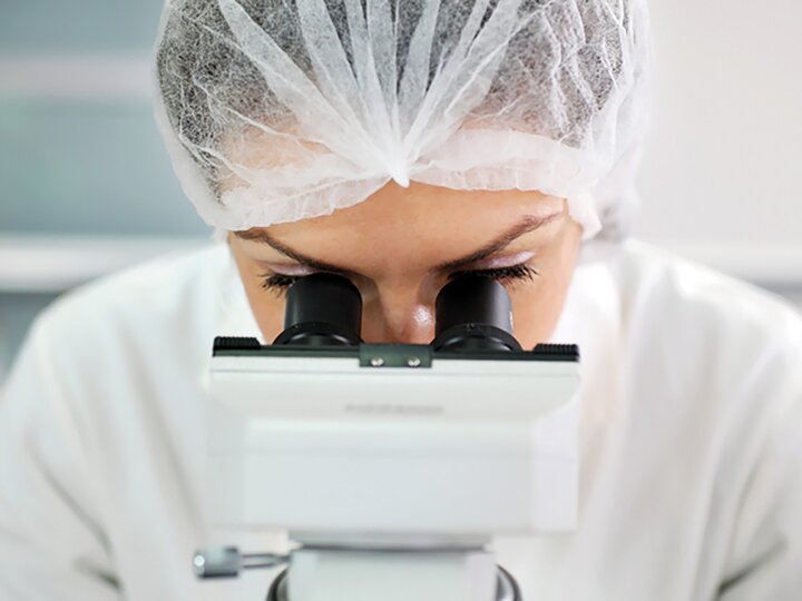 Labormitarbeiterin mit Haarnetz und weißem Kittel schaut in ein weißes Mikroskop