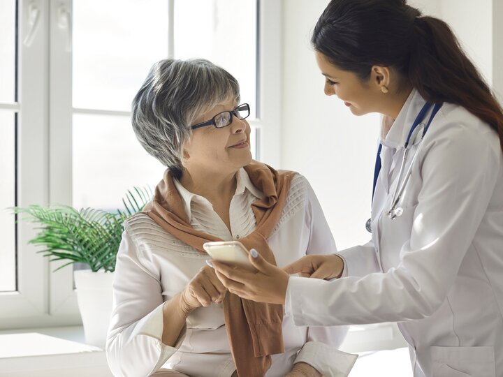 Ärztin mit langen dunklen Haaren in Arztkittel und mit Stethoskop zeigt Patientin mit grauen kurzen Harren und Brille etwas am Smartphone