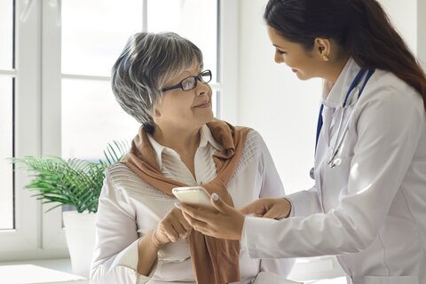 Ärztin mit langen dunklen Haaren in Arztkittel und mit Stethoskop zeigt Patientin mit grauen kurzen Harren und Brille etwas am Smartphone