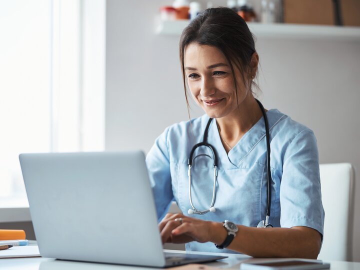 Junge Ärztin mit dunklen Haaren und blauer Arztkleidung sitzt lächelnd am Schreibtisch und schreibt an einem Laptop.