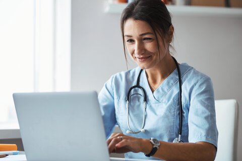 Junge Ärztin mit dunklen Haaren und blauer Arztkleidung sitzt lächelnd am Schreibtisch und schreibt an einem Laptop.