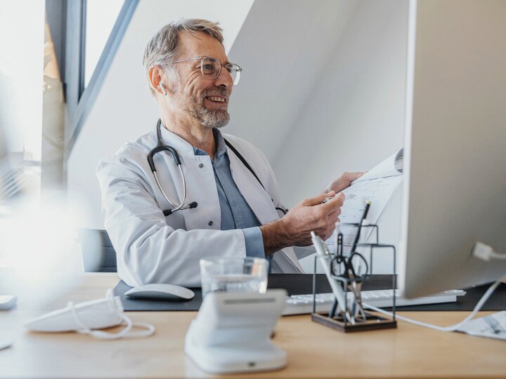Arzt mit grauen Haaren und Brille sitz am Schreibtisch.