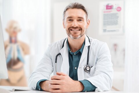 Arzt mit Dreitagebart, Arztkittel und Stethoskop sitzt lächelnd am Schreibtisch. Dahinter verschwommen ein Plakat sowie eine Anatomiefigur.