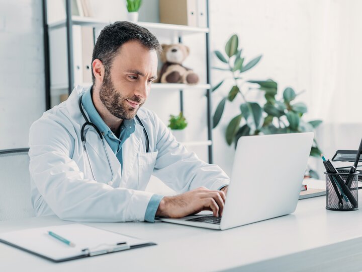 Arzt mit Kittel und Stethoskop sitzt am Schreibtisch und schaut in einen aufgeklappten Laptop. Im Hintergrund verschwommen ein Regal mit Ordnern und einem Teddy.