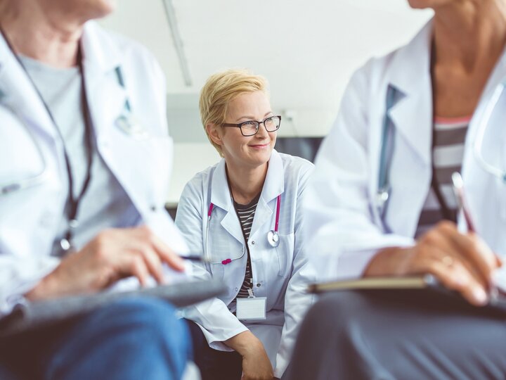 Sitzende Ärztin mit kurzen blonden Haaren. Im Vordergrund unscharf 2 sitzende Ärzte, von denen nur ein Teil des Oberkörpers zu sehen ist.