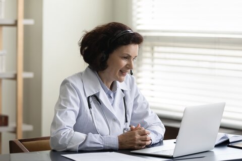 Ärztin sitzt am Schreibtisch. Trägt ein Headset und schaut in einen Laptop.