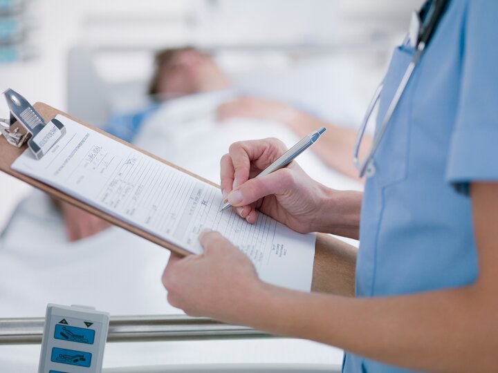 Pfleger füllt Formular auf Klemmbrett aus. Patient im Krankenbett verschwommen im Hintergrund.